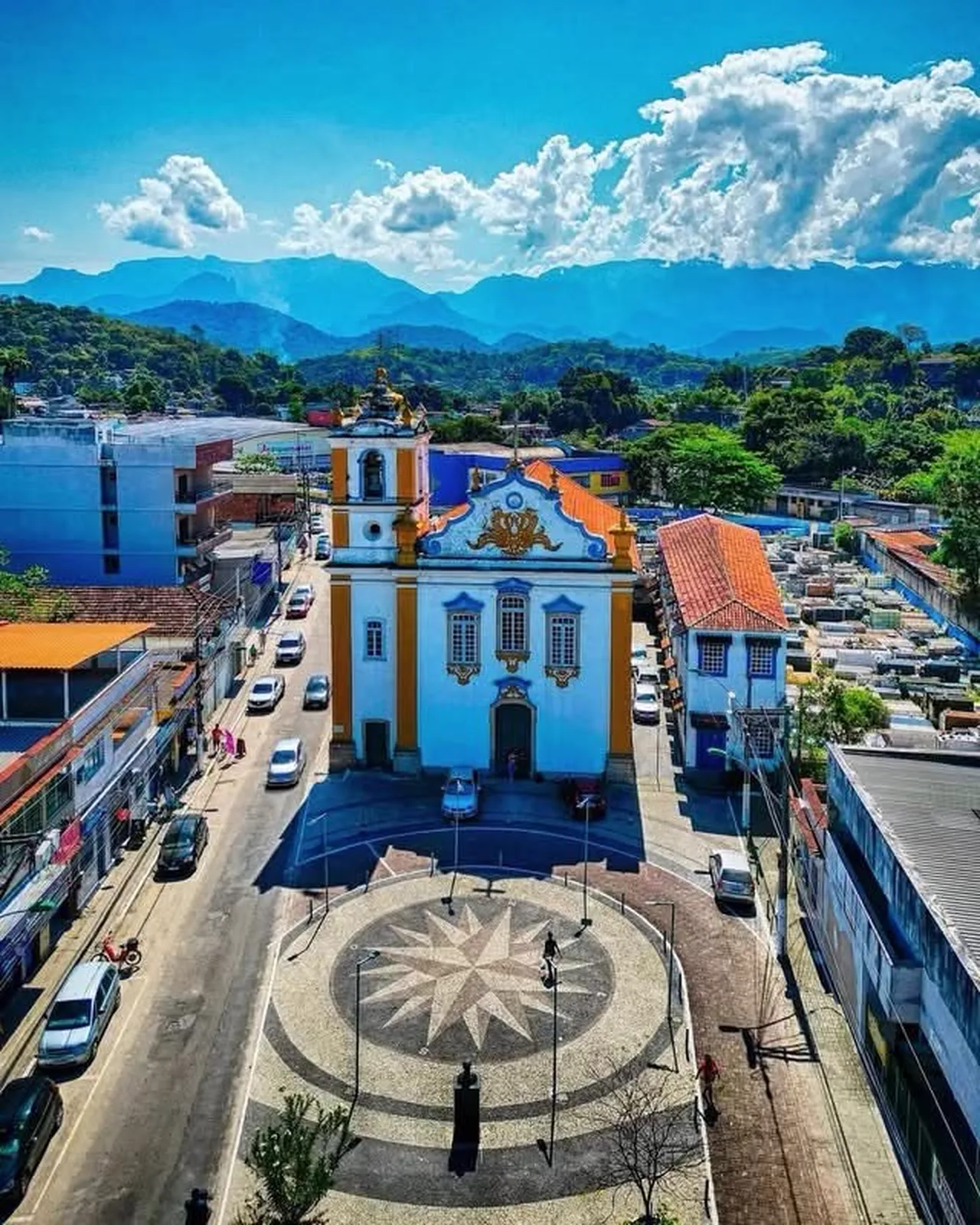 Vista aérea do centro de Magé: a igreja e a praça com mosaico em rosa dos ventos, e a serra ao fundo.